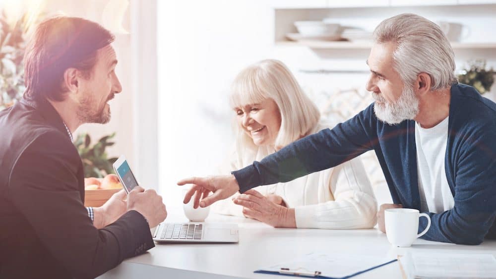 Cheerful professional financial advisor showing the information on laptop while meeting with elderly loving couple in the kitchen