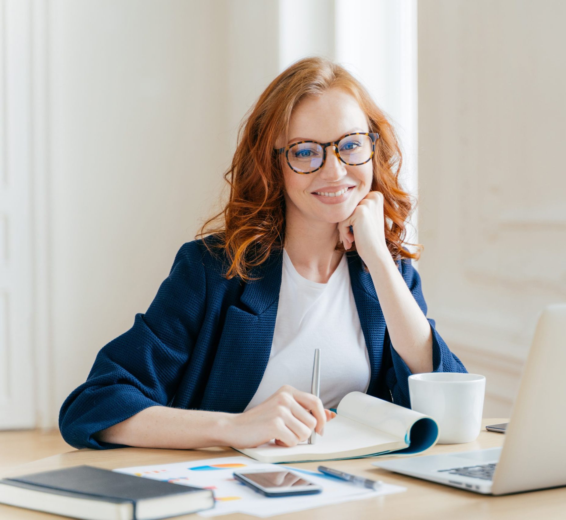 Successful economist in eyewear, checks information on laptop computer, reads information on financial website, writes in notepad and drink hot coffee, sits at workplace. Business and work concept